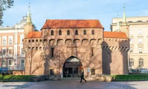 Krakowski barbakan z cegły, z charakterystycznymi wieżyczkami i łukowym przejściem, na tle błękitnego nieba.