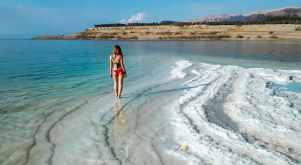 Kobieta w czerwonym bikini spaceruje po krystalicznej plaży Morza Martwego, doświadczając jego niezwykłego zasolenia.