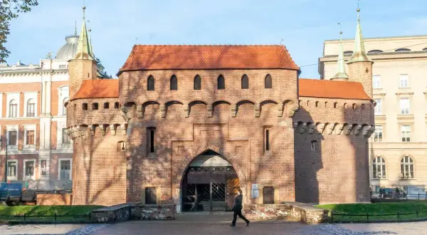 Krakowski barbakan z cegły, z charakterystycznymi wieżyczkami i łukowym przejściem, na tle błękitnego nieba.