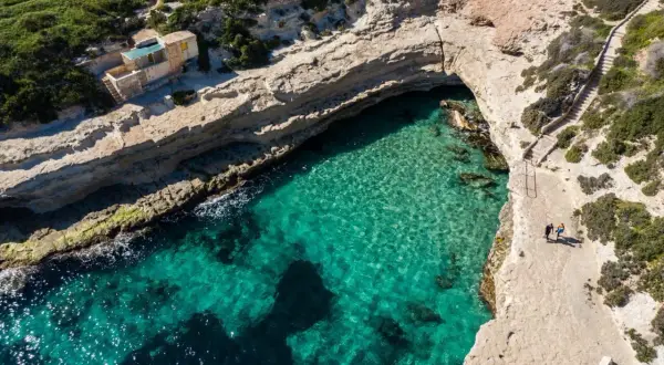 Malta plaze: turkusowa woda, skaliste wybrzeże i mały domek nad zatoczką. Dwie osoby spacerują ścieżką.
