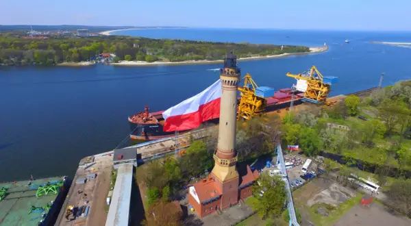 Najwyższa latarnia morska w Polsce dumnie prezentuje biało-czerwoną flagę. Obok cumuje statek, a w tle widać port i morze.