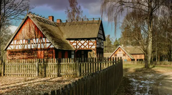 Drewniane domy z krytymi strzechą dachami i drewnianym płotem na tle drzew.