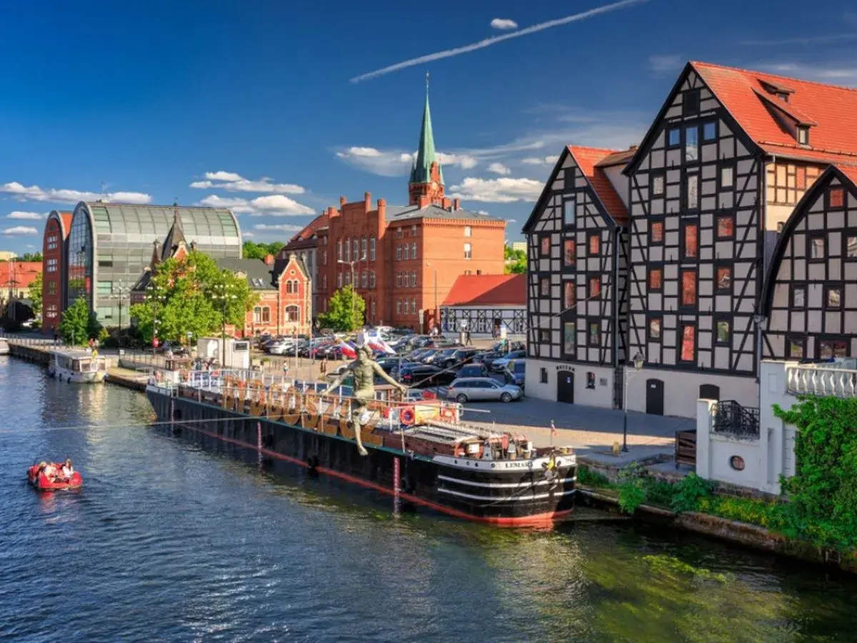 Nad rzeką w Bydgoszczy, jednym z największych miast w Polsce, cumuje statek. W tle zabytkowe kamienice i nowoczesna architektura.