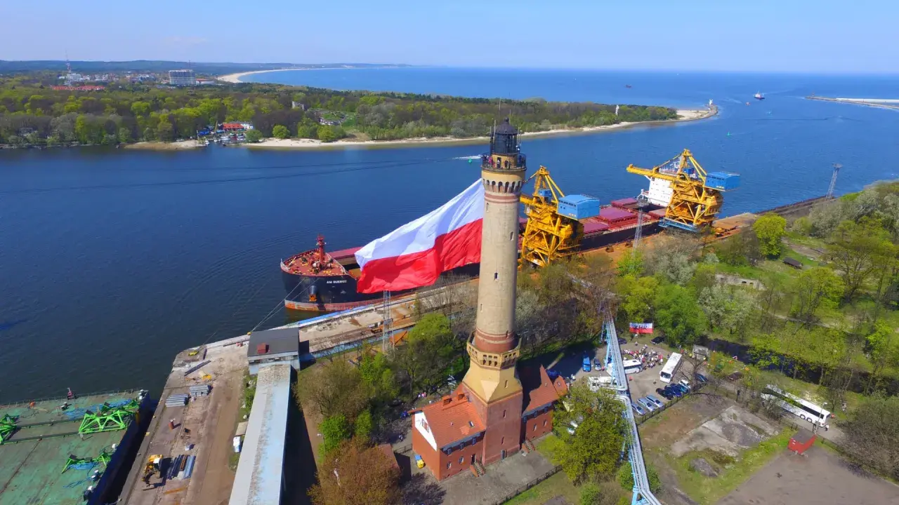 Najwyższa latarnia morska w Polsce dumnie prezentuje biało-czerwoną flagę. Obok cumuje statek, a w tle widać port i morze.