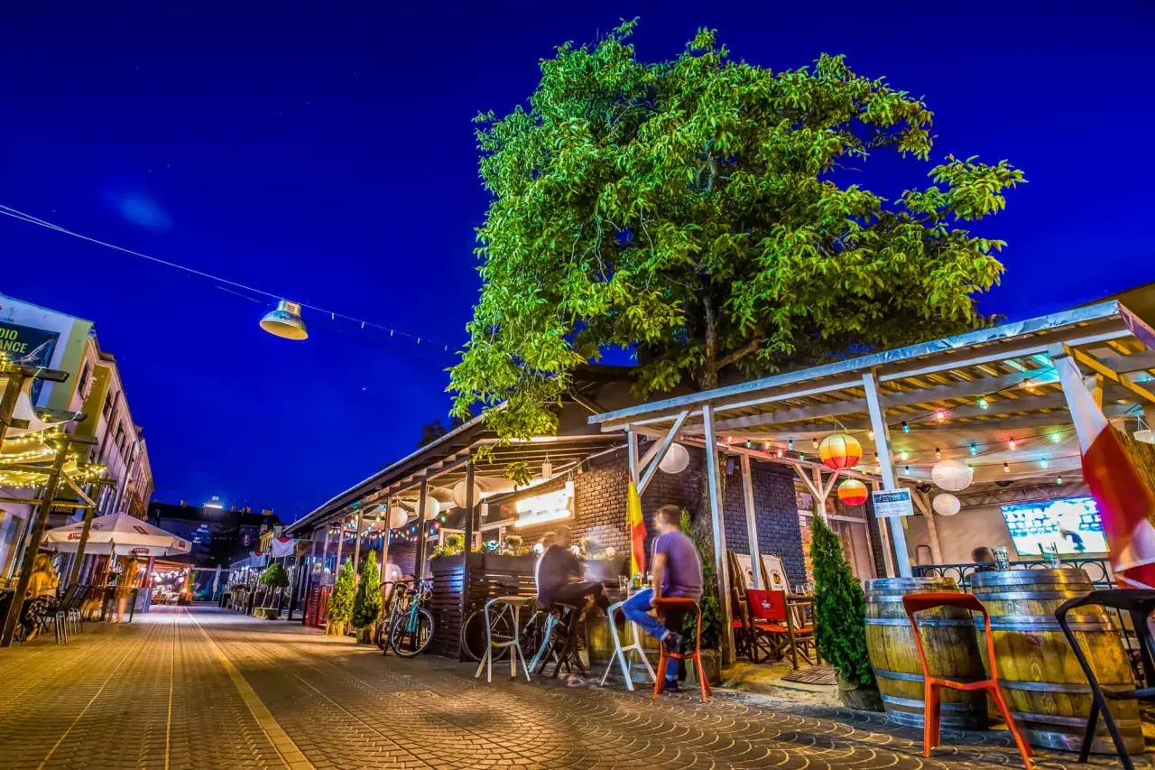 Wieczorna ulica z ogródkami restauracyjnymi, drzewem i oświetleniem.