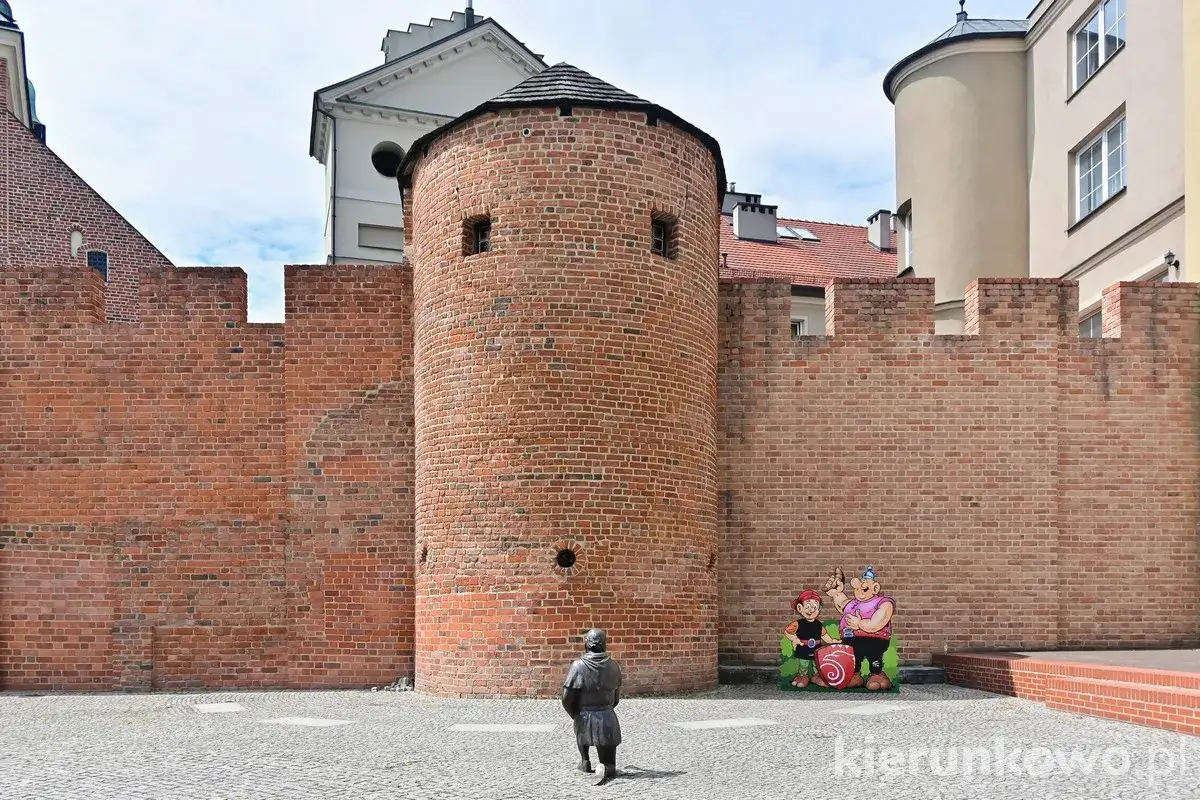 Kamienna wieża i mury obronne, świadkowie historii najstarszego miasta w Polsce. Obok figurki postaci z bajek.