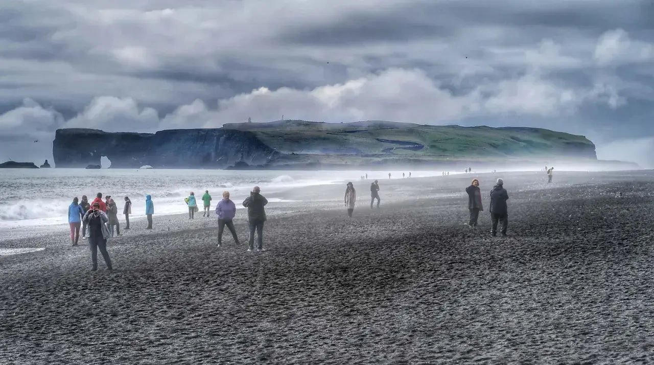 Ludzie spacerują po czarnej plaży pod pochmurnym niebem. W oddali widać klify i skalne łuki.